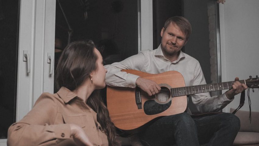 Man and woman playing guitar and drum together in a cozy indoor setting near a window.